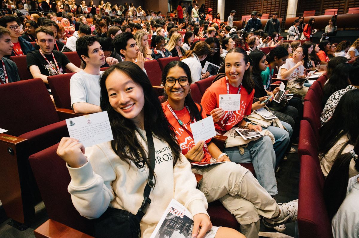 Three smiling first-year students hold up the handwritten messages they received, with dozens of students in the auditorium background. 