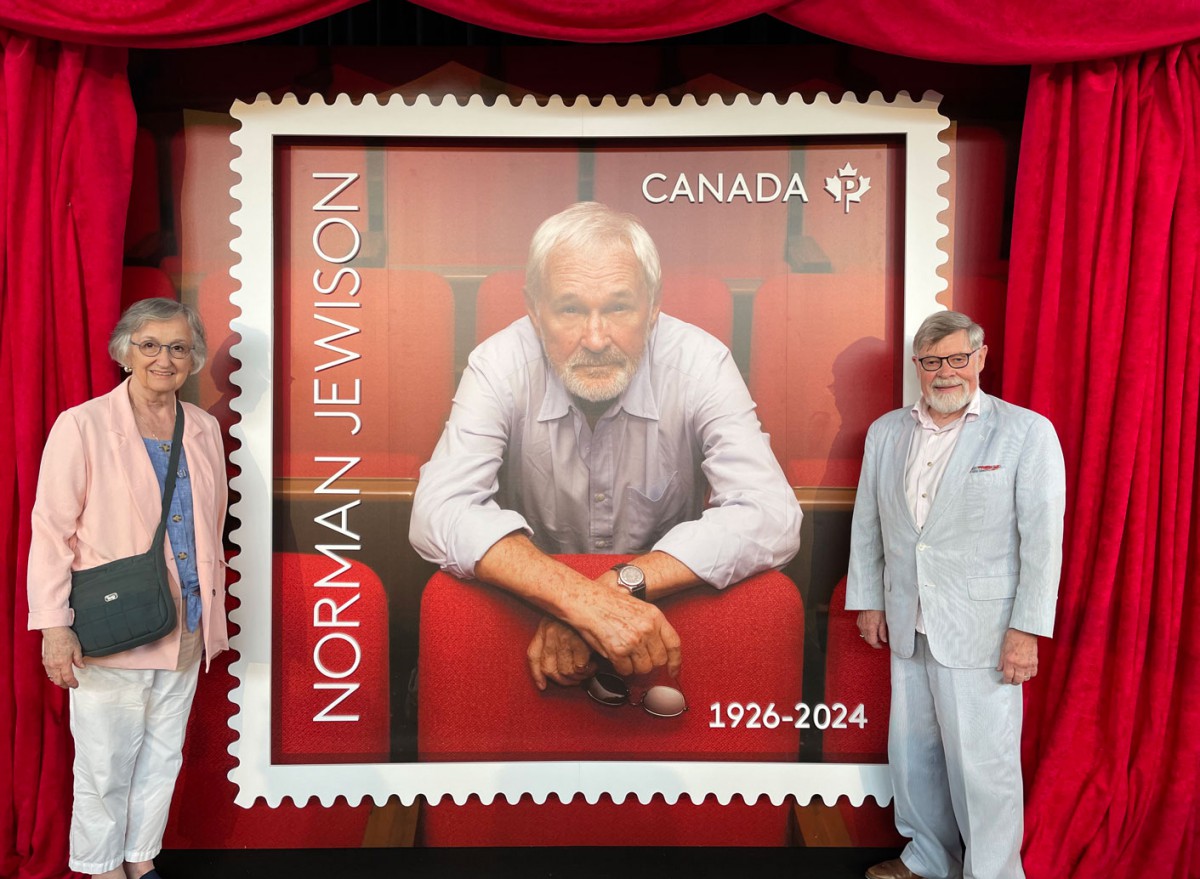 Pauline Thompson and Paul Gooch stand on either side of a large display version of the Norman Jewison commemorative stamp, smiling proudly.